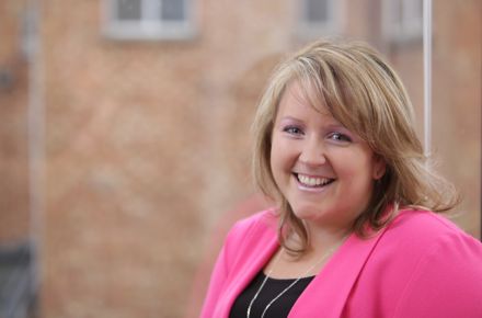 A Kohler Co. employee who works in accounting and finance smiles at the camera. She is wearing a bright pink jacket.