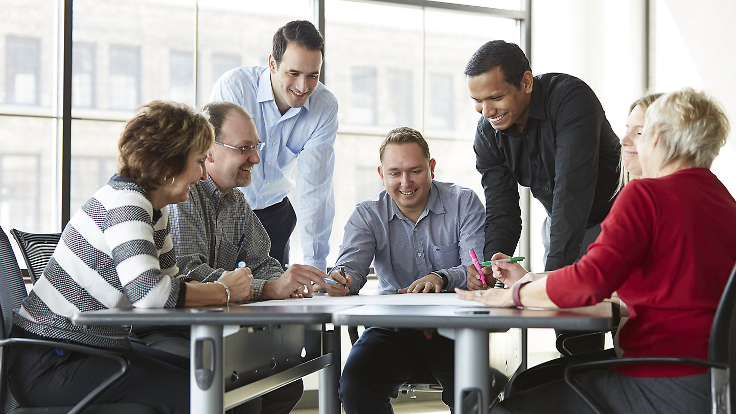 A team of six employees gathers around a table discussing ideas for Kohler Co., each wearing business casual attire