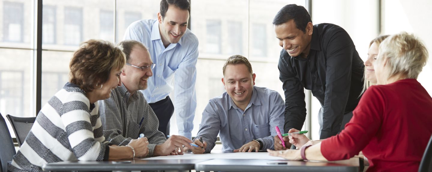 A team of six employees gathers around a table discussing ideas for Kohler Co., each wearing business casual attire