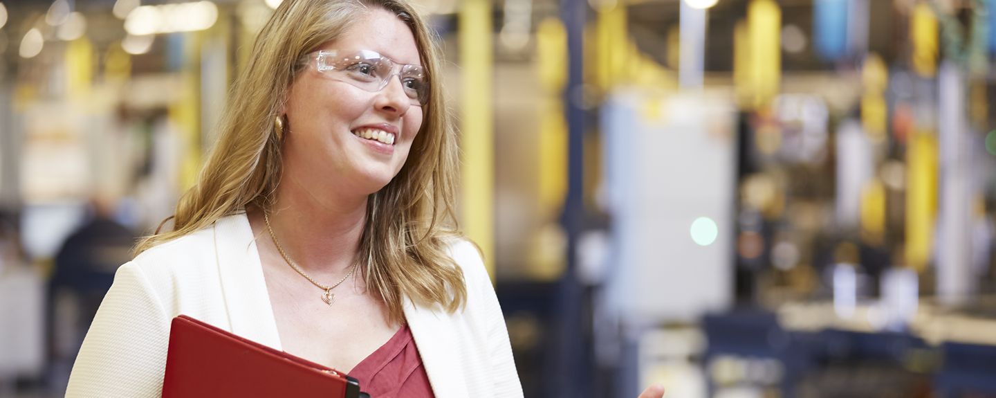 An operations and quality inspector at Kohler Co. stands in a manufacturing facility holding a red binder