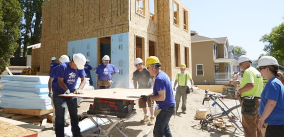 Kohler Power employees gather around a table saw in front of a partially finished home they are building in partnership with Habitat for Humanity