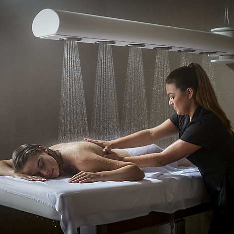 A woman lies on a massage table with a towel draped over her lower half. Water rains down on her as she's massaged by a Kohler Waters Spa employee