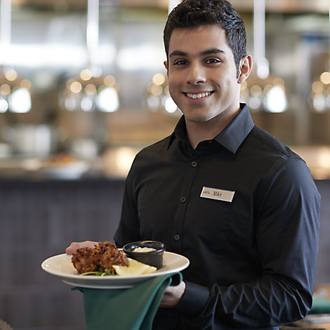 A man with a black polo shirt and name tag stands in the foreground while a Destination Kohler restaurant kitchen appears in soft focus behind him