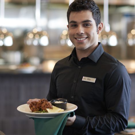 A man with a black polo shirt and name tag stands in the foreground while a Destination Kohler restaurant kitchen appears in soft focus behind him