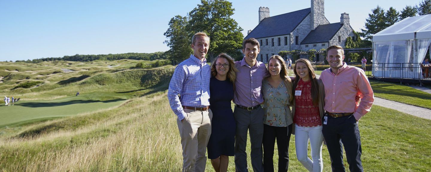 A group stands close together outside with Whistling Straits golf course in the background