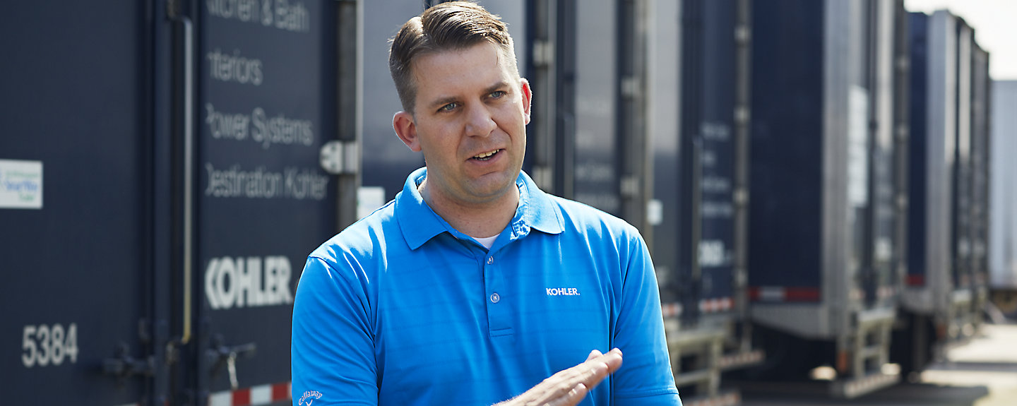 A man wearing a blue Kohler polo shirt stands in front of a row of semi trucks with the Kohler logo.