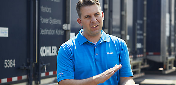 A man wearing a blue Kohler polo shirt stands in front of a row of semi trucks with the Kohler logo on them discussing logistics and supply chain jobs at Kohler Co.