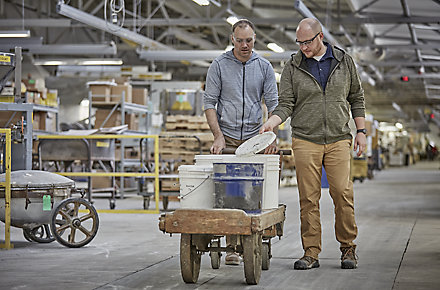 Two employees from Kohler WasteLAB push a cart with five-gallon buckets of manufacturing waste that will be reused