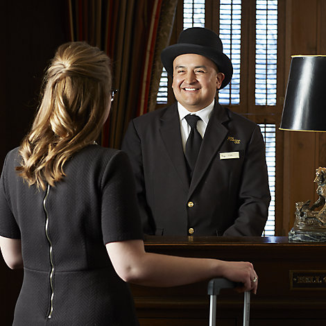 A man wearing a formal hat and suit greets a woman in a black dress in the lobby of The American Club hotel
