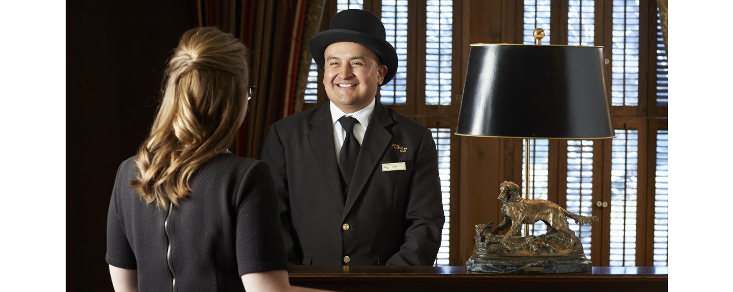 A man wearing a formal hat and suit greets a woman in a black dress in the lobby of The American Club hotel