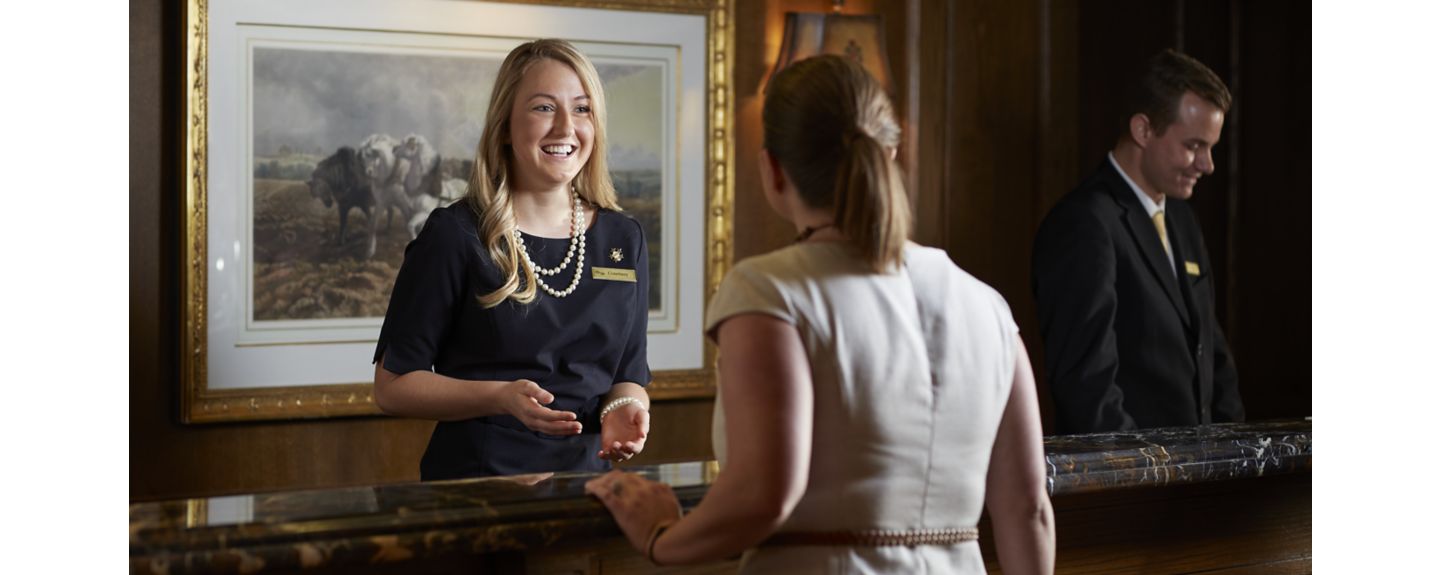 A woman in a black dress and pearls greats a guest at The American Club's front desk. Another employees stands to her right.