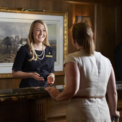 A woman in a black dress and pearls greats a guest at The American Club's front desk. Another employees stands to her right.