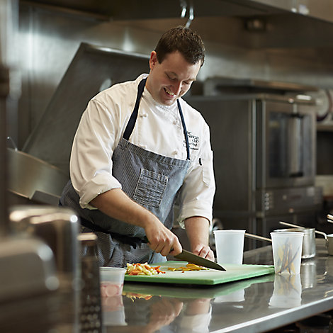 One of the Destination Kohler chefs chops colorful produce on top of a steel kitchen counter while a chef in the background cooks on the stovetop