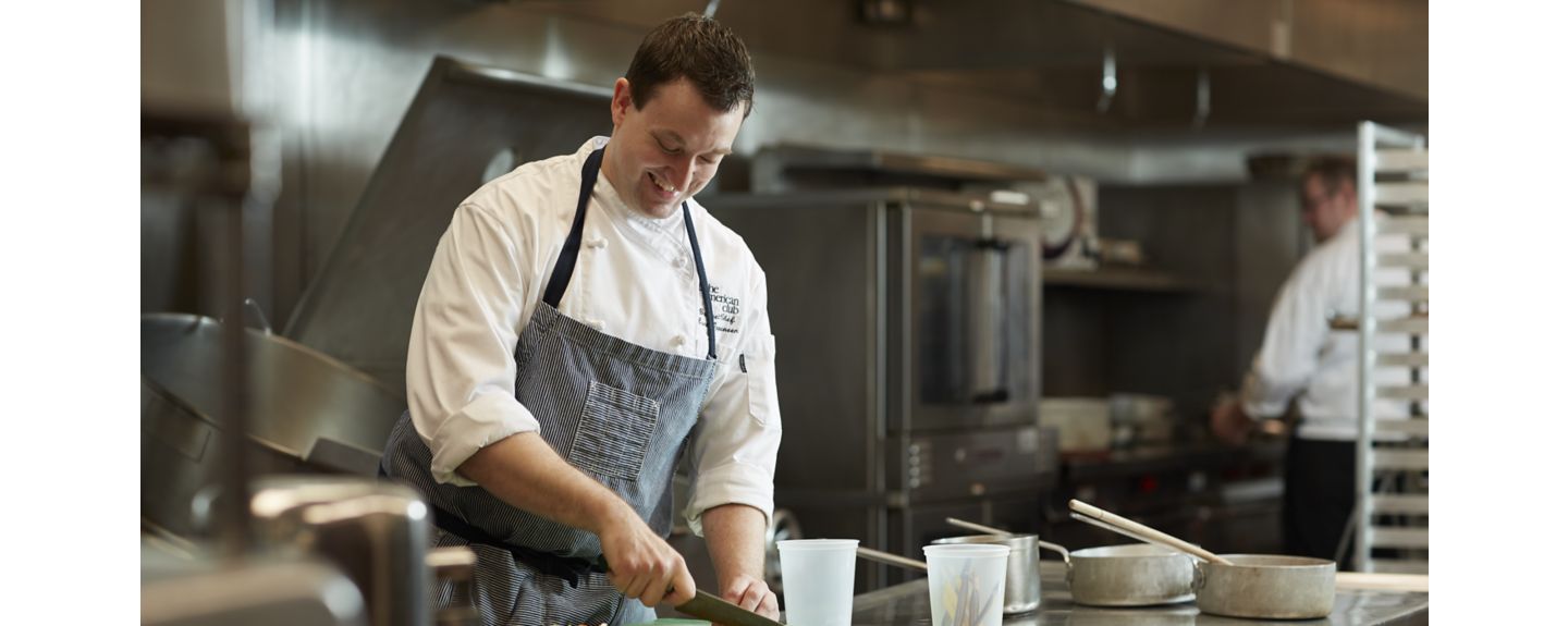 A Destination Kohler chef wearing a chef's coat and apron uses a mandoline to slice an onion in a restaurant kitchen