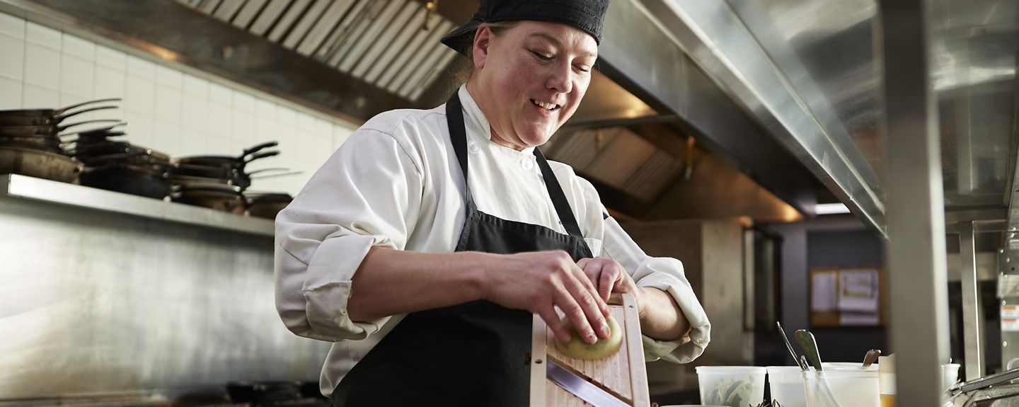 A Destination Kohler chef wearing a chef's coat and apron uses a mandoline to slice an onion in a restaurant kitchen