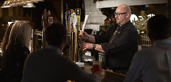 A bartender wearing a black shirt and tie pours a draft beer for guests sitting at the bar in front of him in Kohler, Wisconsin