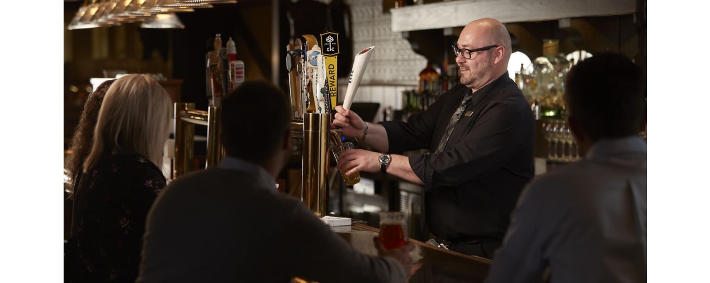 A bartender wearing a black shirt and tie pours a draft beer for guests sitting at the bar in front of him in Kohler, Wisconsin