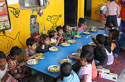 15 children sit around a big table eating with colorful paintings on the wall behind them