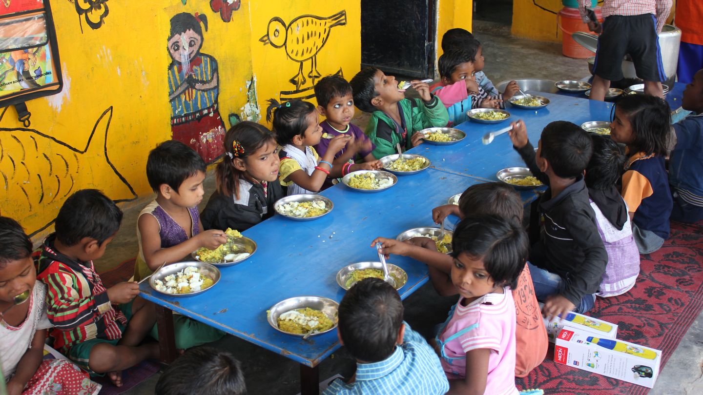 15 children sit around a big table eating with colorful paintings on the wall behind them