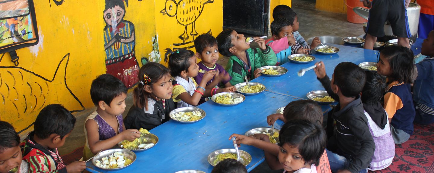 15 children sit around a big table eating with colorful paintings on the wall behind them