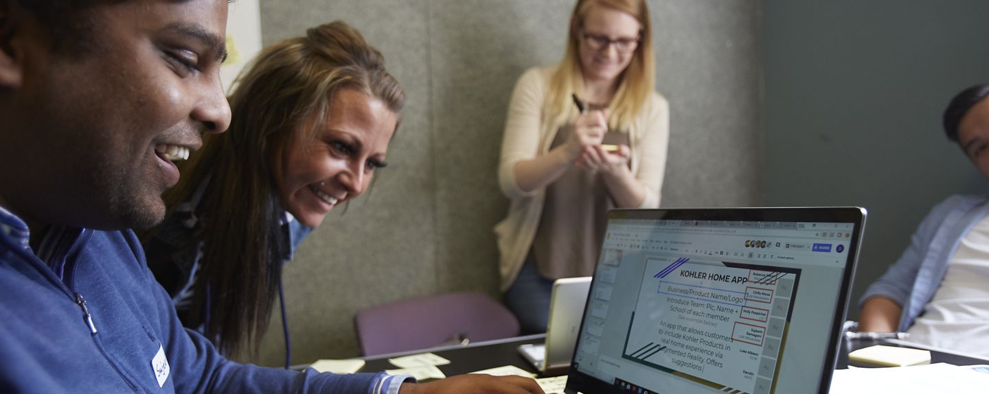 Two Kohler Co. employees look at a laptop together, both smiling, and other employees are in the background.