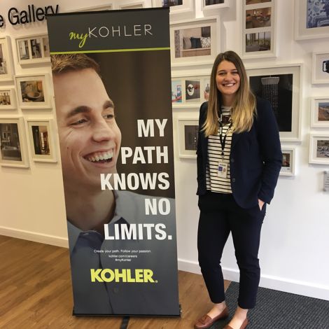 Cristina Bigica, Marketing and Communication – Buyer for Kohler Co. in Gloucester, UK, stands in front of a wall of framed artwork next to a sign that says My Path Knows No Limits