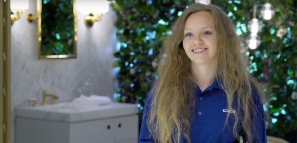 A woman in a blue polo shirt with Kohler logo smiles at the camera. A Kohler vanity and plants are in the background