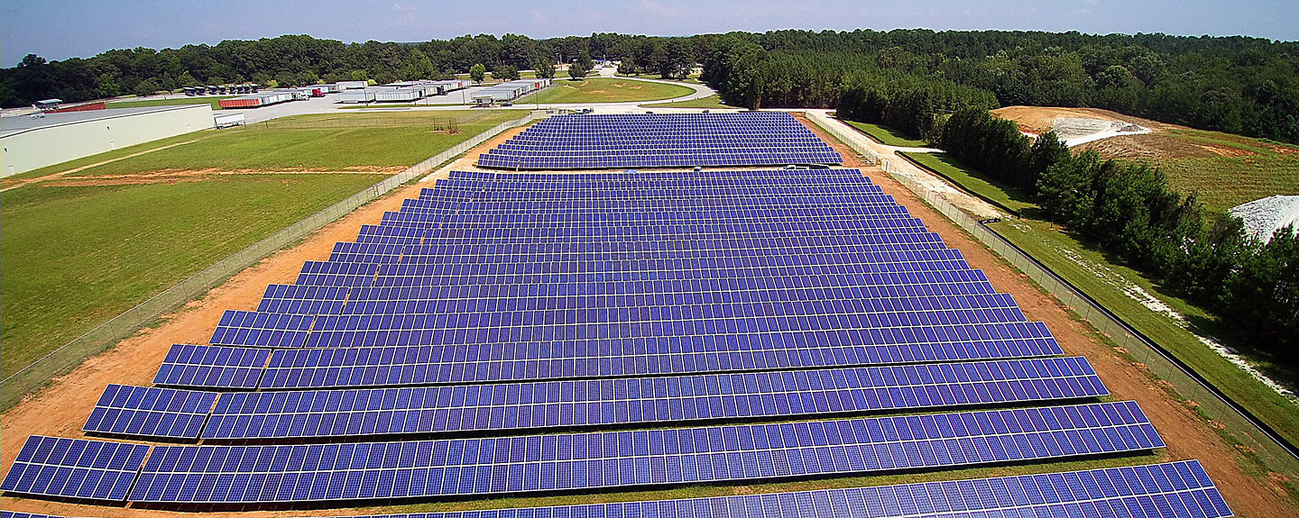 An aerial view of rows of solar panels next to a field, trees, and a factory