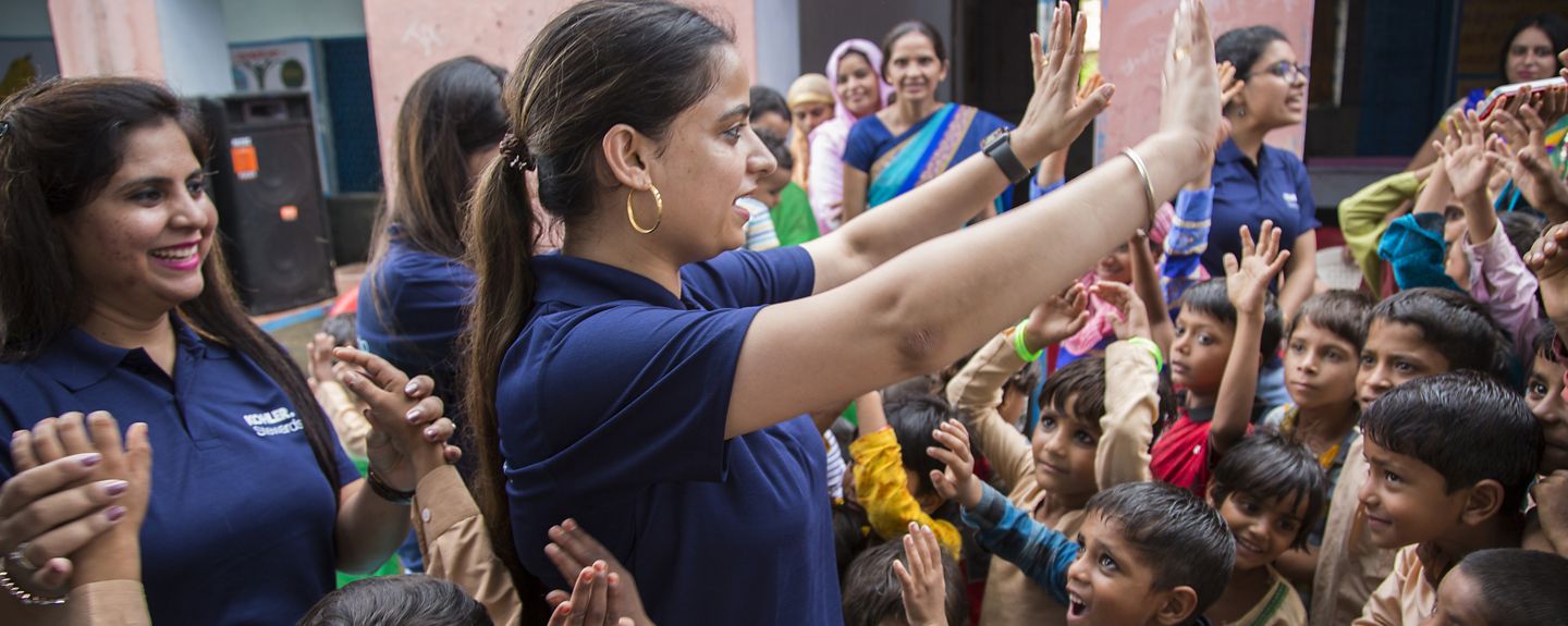 A large group gathers as one woman, raises her hands while talking to children.