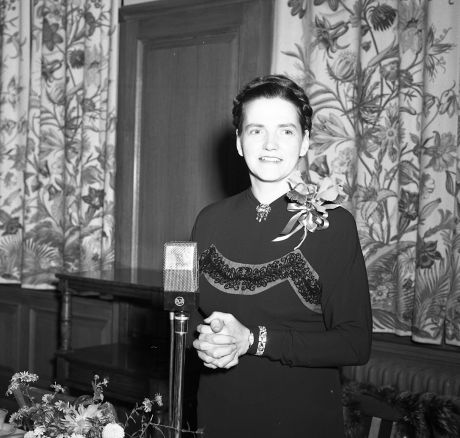 Ruth Kohler stands in front of a microphone and decorative floral tapestries in this black and white photo