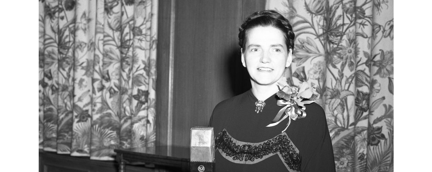 Ruth Kohler stands in front of a microphone and decorative floral tapestries in this black and white photo