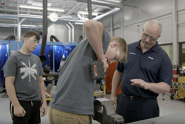 A teenage boy, part of Kohler Co.'s Skilled Trades program for high school students, uses a hand drill as another teenage boy and an adult look on