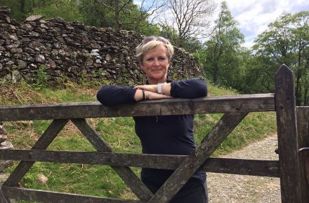 Linda Waite, NPD Project Leader for Kohler Co. in Cheltenham, England, stands behind a fence with her arms crossed. A stone wall and trees are in the background