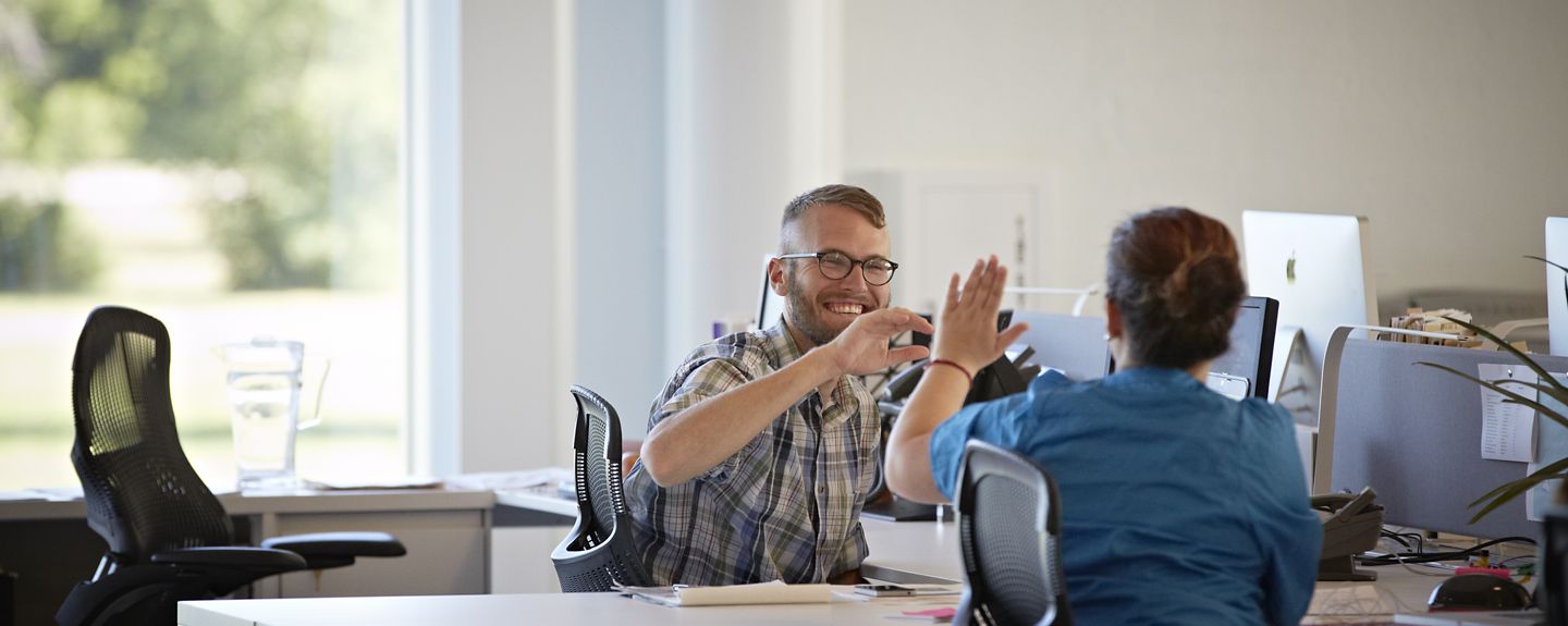 Two employees in Kohler Co.'s marketing and communications department reach over their bookshelves.
