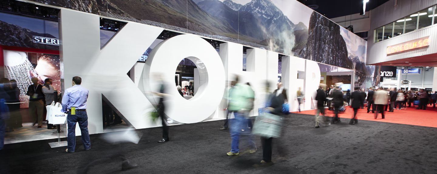 A busy scene shows people walking by a big KOHLER sign at a trade show