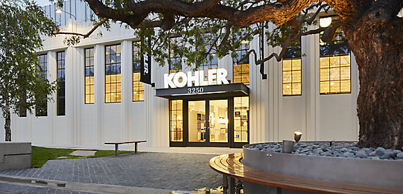 A photo taken from outside shows the front entrance to a LEED-certified KOHLER showroom. The interior is lighted and a KOHLER sign hangs by the front door