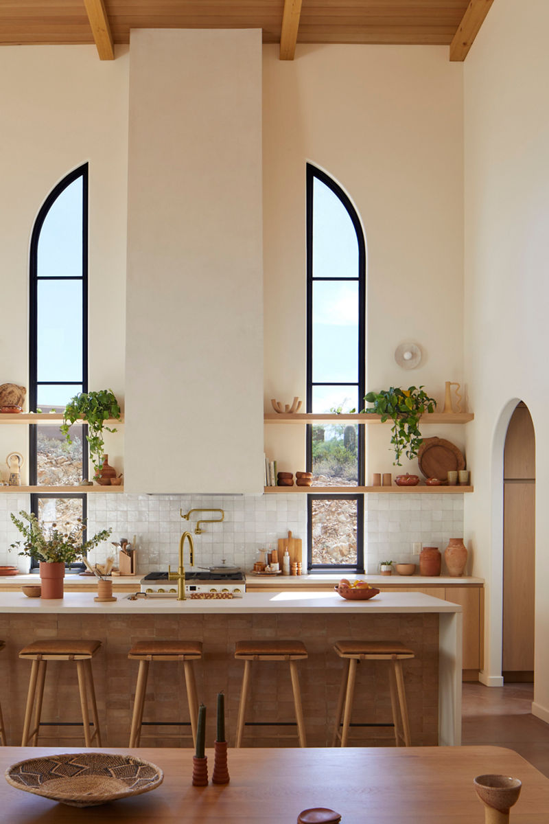 Vertical image of the vaulted ceiling kitchen in Christina and Robert's home. The range hood goes all the way up to the ceiling with two symmetrical, tall windows on each side. Under the range hood is a Kohler pot filler faucet in brass which matches the brass Kohler kitchen faucet on the kitchen island in the center of the room.  