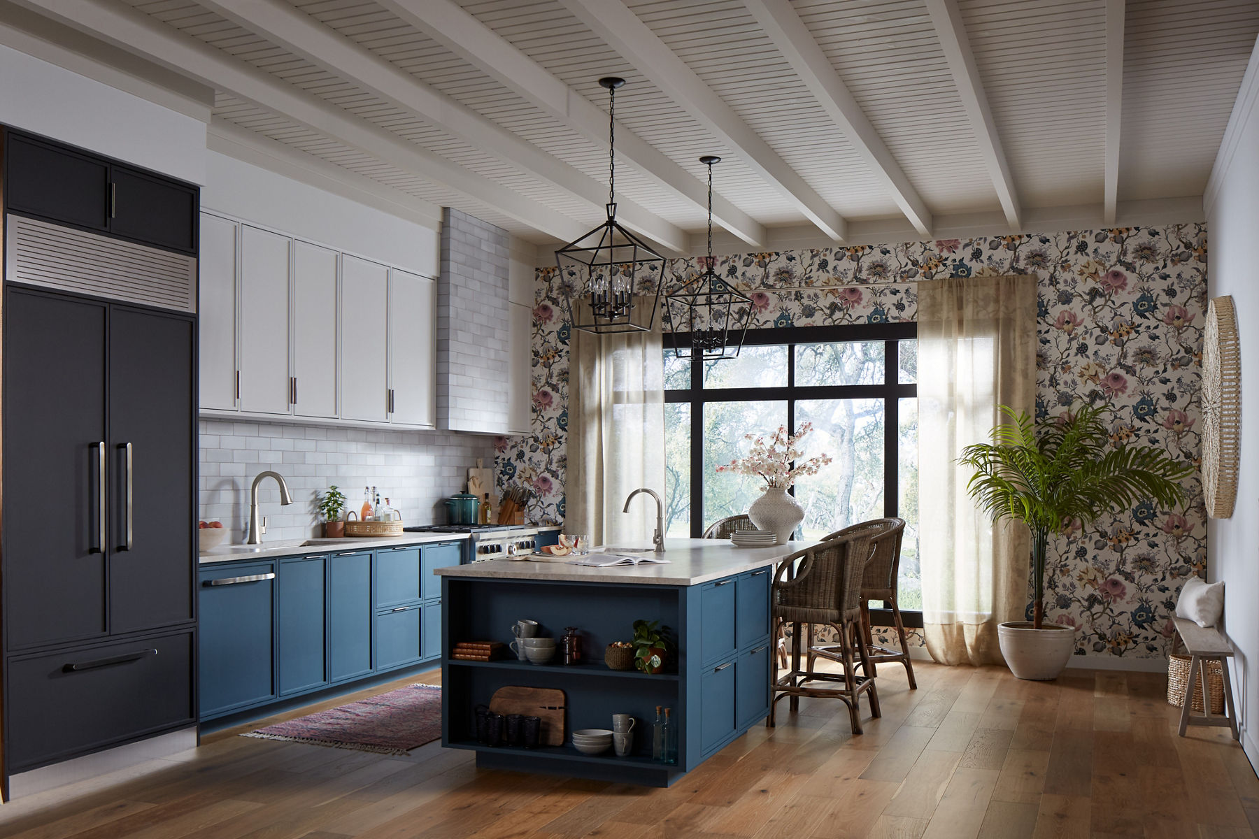 A kitchen with a large blue island with white top, island prep sink with KOHLER single-hole faucet, blue lower cabinets with white uppers, Riff faucet, and one wall with blue and white floral wallpaper.