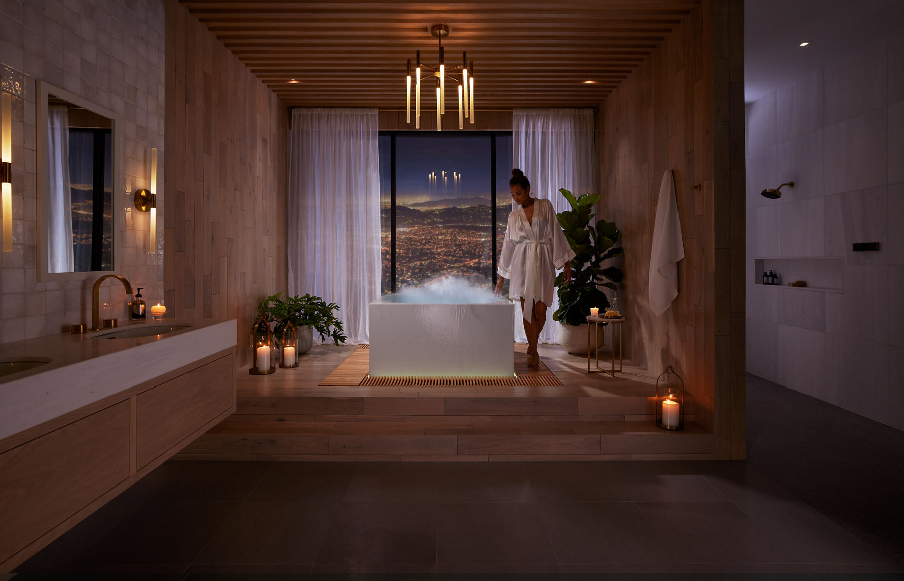 Une grande salle de bain avec une femme au centre marchant le long du côté d’une baignoire Stillness, avec de la vapeur qui s’élève de la surface, avec une grande fenêtre en arrière-plan surplombant un paysage urbain nocturne et des montagnes au loin.