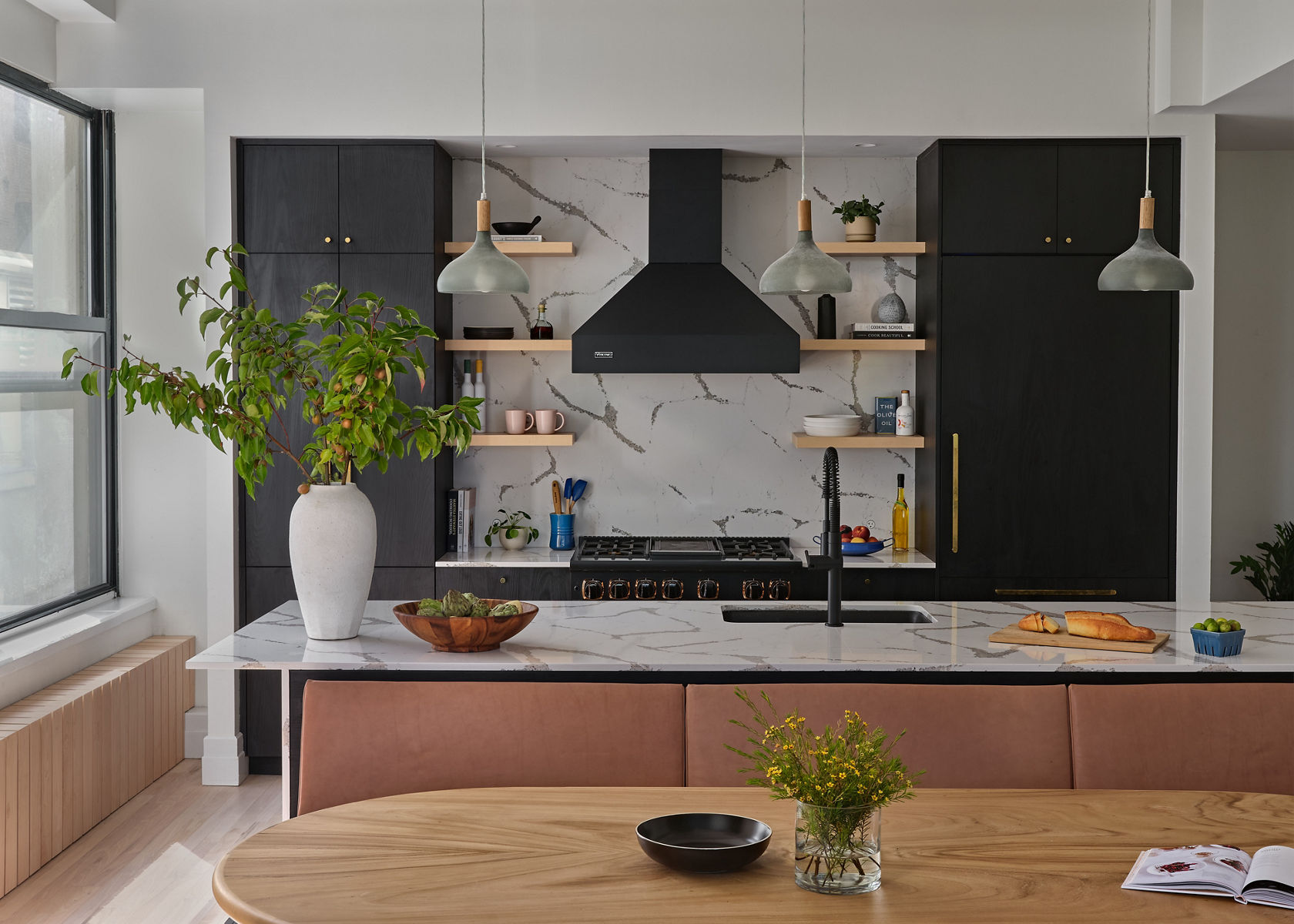 An open kitchen in Japandi style features an oval table with bench seating, an island with KOHLER Crue Semi-Professional faucet, and black floor to ceiling cabinets flanking a marble wall with range hood, wood open shelving, and a professional range.