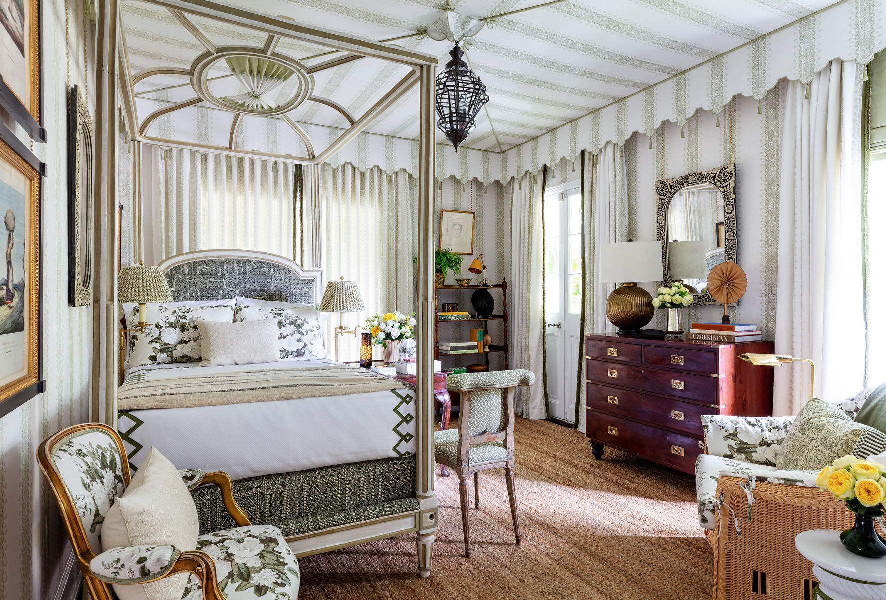 A Grandmillennial style bedroom with green and white striped wallpaper, canopy bed, bamboo display shelf, and an assortment of chairs in contrasting styles and upholstery patterns.