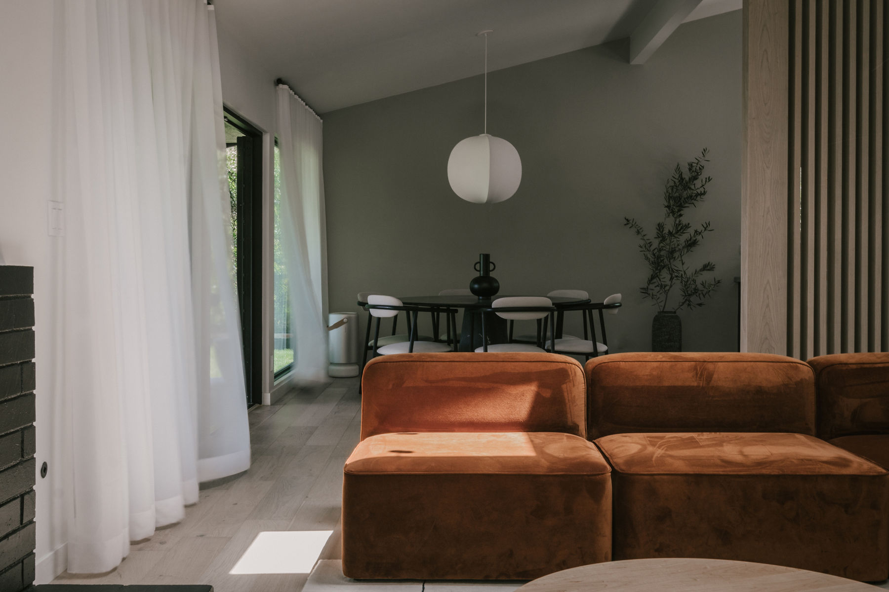 An open plan home in Japandi style features white curtains over floor to ceiling windows, light wood floors, a rust colored sofa, a black dining table with white chairs, a white pendannt light, and sage green walls.