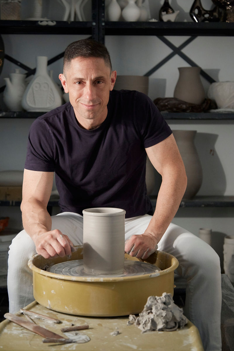 Portrait of designer Jonathan Adler at his pottery wheel, looking into the camera with vases that he's created behind him on shelves and a half-created vase in front of him, symbolizing his pottery background and creative spirit. 