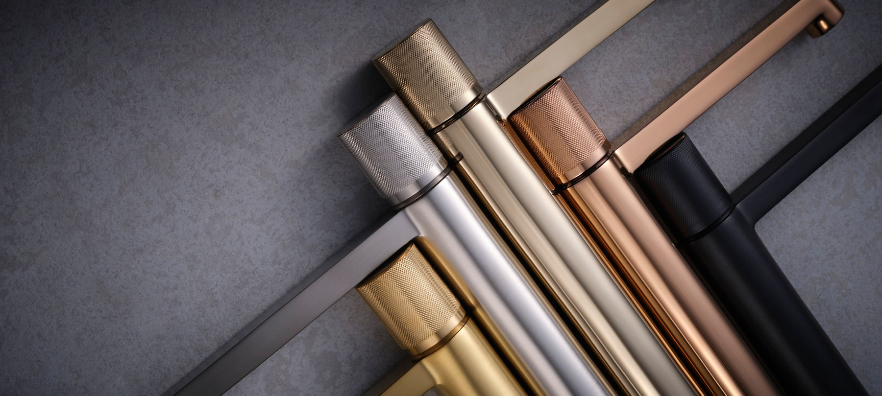 Close-up view of various metallic faucet handles in different finishes, including brushed nickel, gold, bronze, and stainless steel. The handles feature a textured, knurled design, highlighting their craftsmanship and variety of color options.
