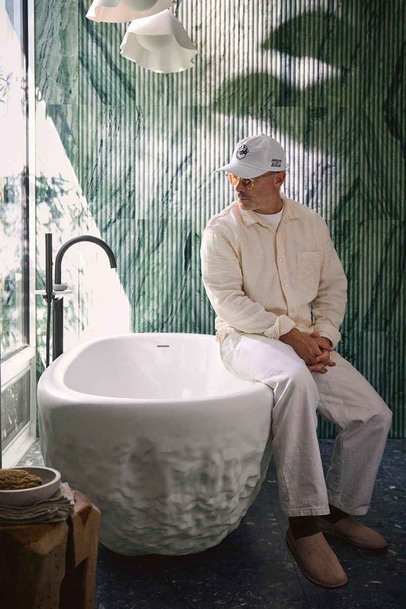 Designer seated on the edge of a sculptural freestanding bathtub in a modern bathroom with green textured walls. The designer is wearing a light-colored outfit and a cap, gazing thoughtfully to the side.
