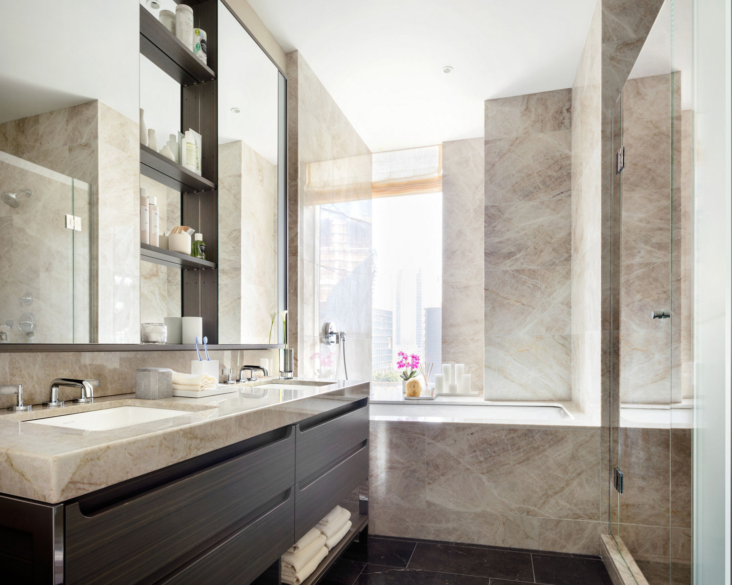 Interior view of a bathroom of One Hudson Yards featuring light brown marble on the walls and countertop with undermount sinks and bath under a large mirror reflecting the large glass-enclosed shower. 