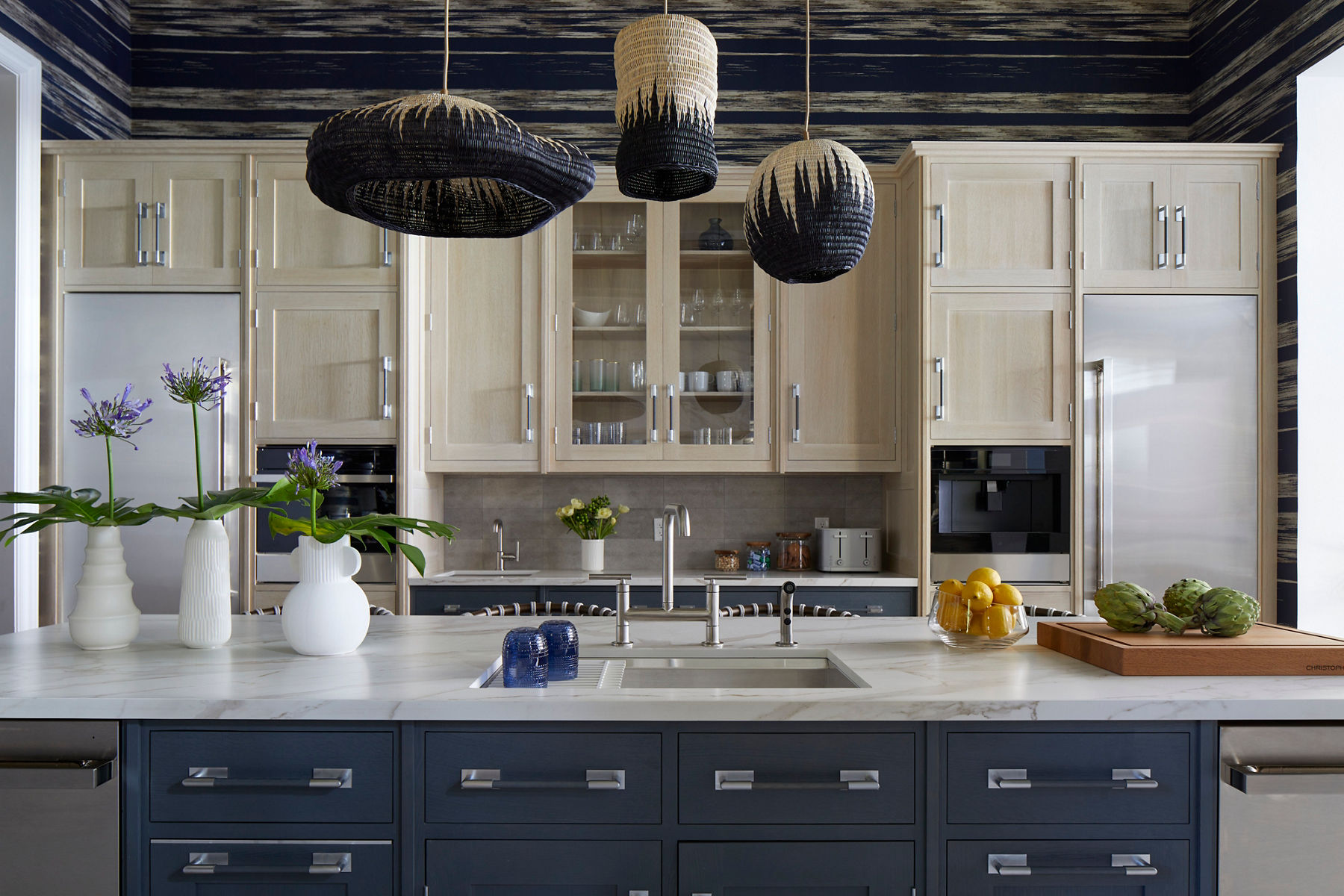 Wide image of a large kitchen island and wall of cabinets behind it. At the center of the island is a sink with faucet centerpiece. The base cabinets are colonial blue with the upper cabinets in cream. A soothing patterned wallcovering is applied above the cabinets.