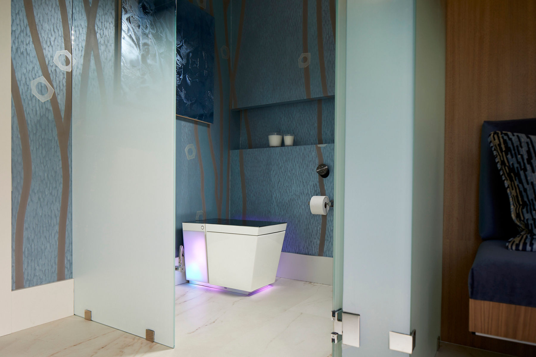 Image of a white smart toilet with a glowing light at its base. The frosted-glass door of the toileting space is open with frosted-glass panels along the side for privacy. The blue walls feature a texture and tree-limb design elements.