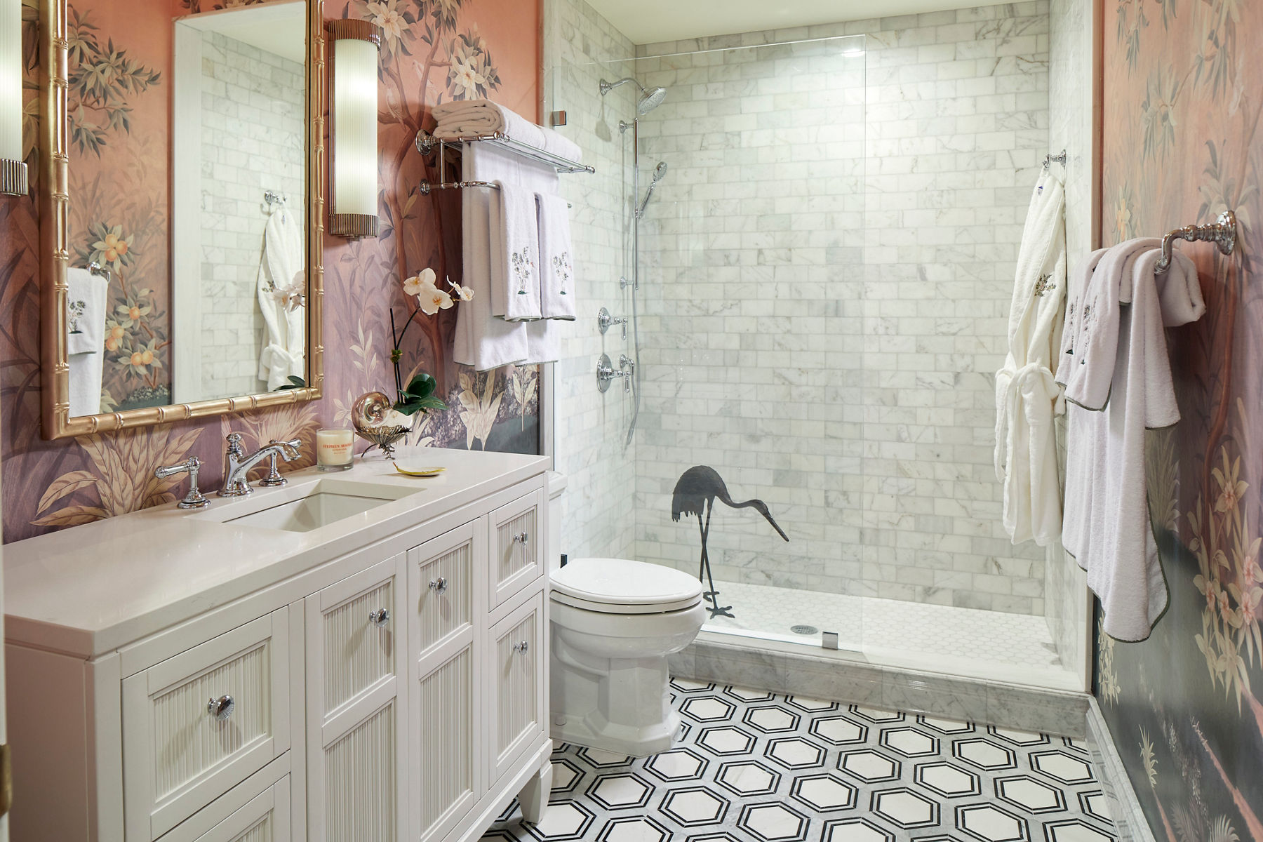 Image of a white bathroom vanity, countertop with square sink, white toilet, and glass enclosed alcove shower, featuring a bird stencil on the base of the glass shower door. Grey patterned tile on the floor complements the tile shower walls, next to a floral pattern wall covering surrounding the vanity mirror.