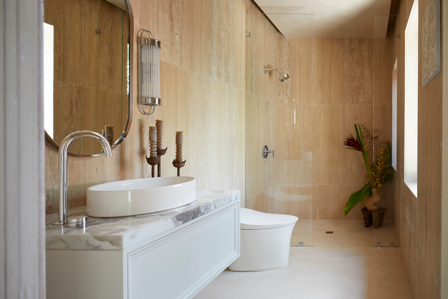 A norrow bathroom with a white and marble wall-hung vanity, oval mirrors, lighting sconces, white toilet, and glass-enclosed shower along one wall, opposite a window on the other wall.